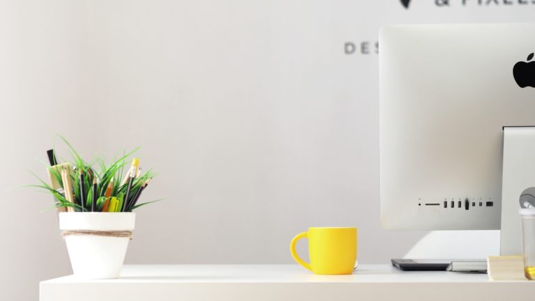 A desk with a computer monitor, bright yellow mug, and planter.