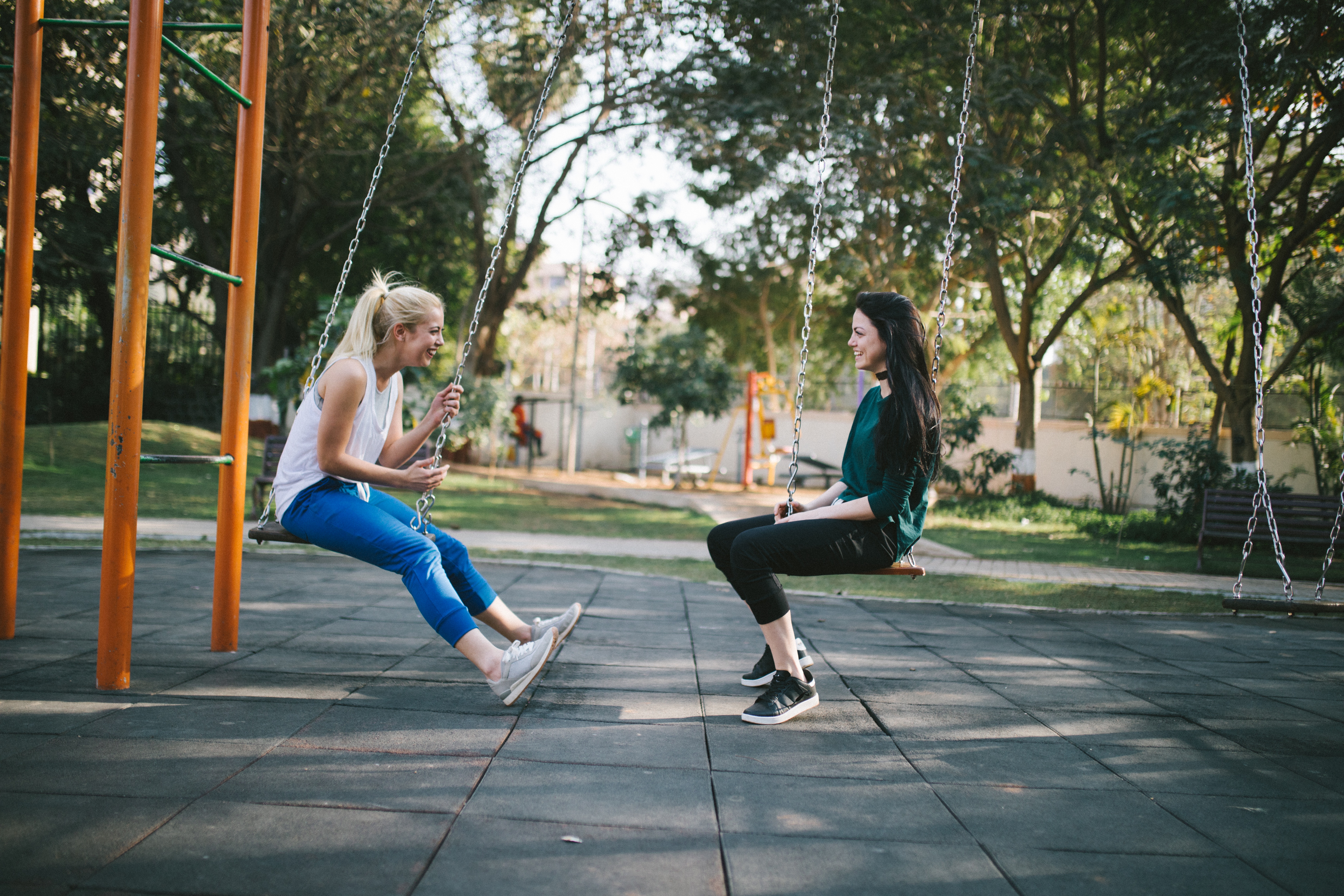 Two friends sitting on swings at a park, laughing and facing each other.