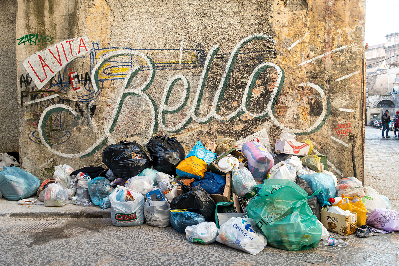 A street corner featuring a "La vita bella" mural and a pile of trash bags.